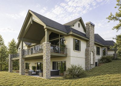 Two-story home with stacked stone pillars supporting a covered deck and lower patio area, stone chimney visible on the right side