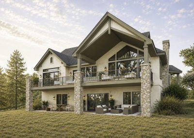 Contemporary two-story home with stone chimney pillars, covered rear deck with metal railing, expansive windows, and manicured lawn sloping downward with evergreen trees in background.