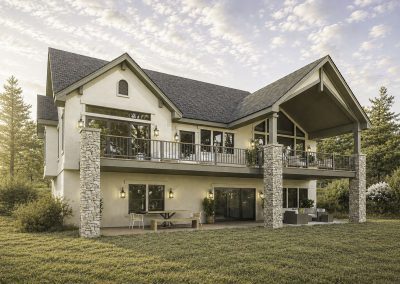 Modern two-story home with white siding, dark roof, and stone support columns framing an expansive rear deck with metal railings overlooking mountain terrain